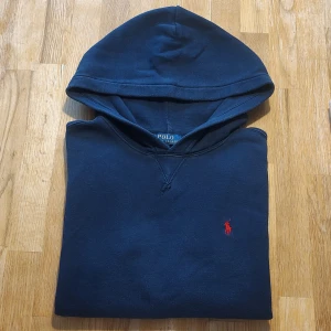 Mörkblå hoodie från Ralph Lauren - Klassisk hoodie från Ralph Lauren, marinblå med vinröd logga. Stl L=14-16, uppskattas dock vara ungefär storlek 152/158, se mått. Fint begagnat skick, enda anmärkning är små svagt blekta partier i trekanten vid halsen.  Mått: Armhåla till armhåla 50cm, bredd längst ner 45cm, längd från mitt på axeln 57,5cm.