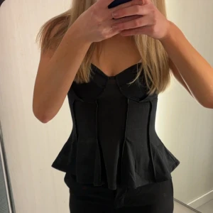 Svart korsett-topp - Snygg svart korsett-topp med en elegant design. Toppen har en figurnära passform och är perfekt för en stilren look. Den är axelbandslös och har en strukturerad form som framhäver figuren.