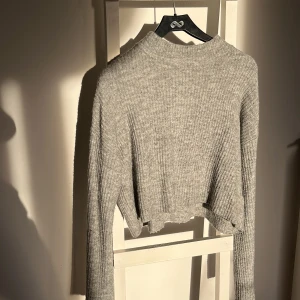 Grå ribbad croppad tröja - Säljer en stilren grå ribbad tröja med croppad passform. Tröjan har långa ärmar och en lite högre hals. Enkel och snygg design som passar till det mesta. Perfekt för dig som gillar en minimalistisk stil.