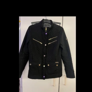 Svart quiltad jacka från Rock&blue - Snygg svart quiltad jacka med dragkedja och knappar framtill. Jackan har flera praktiska fickor med dragkedjor och knappar, samt en stilren design. Perfekt för kyligare dagar.