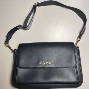 Svart axelväska från Tommy Hilfiger - Snygg svart axelväska från Tommy Hilfiger i skinn med justerbar axelrem och guldfärgade detaljer. Väskan har en stilren design med ett praktiskt magnetlås och flera fack inuti för enkel organisering. Perfekt för att bära dina nödvändigheter med stil.