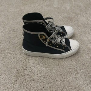Dior high sneakers - Snygga svarta sneakers från Christian Dior med vita sulor och textdetaljer på snörningen. Skorna har en unik design med stjärnmönster på sulorna och guldfärgade detaljer vid snörningen. Även defekter på bla sulan och inne vid hälen men har täckt över med shoe white cream🤍🤍🤍🤍🖤🖤🖤🖤