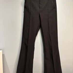 Svarta kostymbyxor - Snygga svarta kostymbyxor med låg midja och bootcut-stil. Perfekta för en stilren look. Byxorna har en klassisk design med en slät front och diskreta sömmar.
