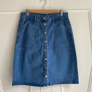 Blå jeanskjol med knappar - Snygg blå jeanskjol med knappar hela vägen fram och praktiska fickor både fram och bak. Kjolen har en klassisk design med bälteshällor i en denim utan stretch. Passar bra till en enkel t-shirt eller en stickad tröja. Midja 79 cm, längd 62. Står storlek L men är nog snarare S/XS.  Aldrig använd. 