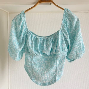 Blommig offshoulder topp i ljusblått oanvänd L - Charmig ljusblå offshoulder topp med blommigt mönster. Toppen har korta ärmar och en volangdetalj vid halsringningen. Perfekt för en somrig look!