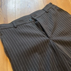 Svart randiga kostymbyxor - Snygga svart kostymbyxor med vita ränder. Byxorna har en klassisk design med dragkedja och knapp framtill. Perfekta för en stilren look.