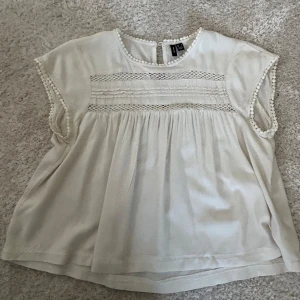 Vit blus från Vero Moda - Super söt vit blus från Vero Moda med spetsdetaljer och korta ärmar. Blusen har en luftig passform och dekorativa spetsar vid ärmslut och över bröstet. Perfekt för en avslappnad och stilren look.