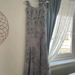 Ljuslila långklänning med blommigt mönster - Elegant ljuslila långklänning med vackra blommiga broderier i grönt och grått. Klänningen har en v-ringad design och axelband som ger en romantisk känsla. Perfekt för speciella tillfällen. (Den är skäddarsydd) storlek: S/M