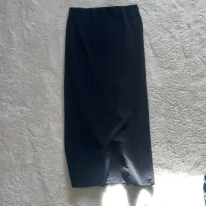 Elegant svart långkjol med en stilren design. Kjolen har en diskret slits baktill som ger extra rörelsefrihet. Perfekt för en chic och minimalistisk look.