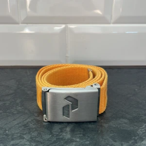 Gult bälte från peak performance  - Snyggt gult bälte från peak performance med ett stilrent silverfärgat spänne. Perfekt för att ge en färgklick till din outfit. Bältet är 105 cm och är otroligt snyggt och stiligt. Även så har bältet en enkel desgin och passar in i de flesta stilarna!