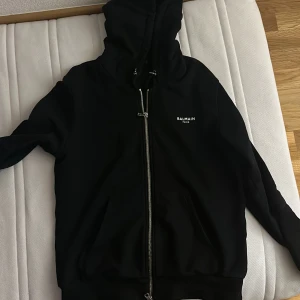 Svart hoodie från Balmain - Säljer en snygg svart hoodie från Balmain. Den har en dragkedja framtill och en huva med snörning. Perfekt för en casual look eller att mysa i hemma. Passar till alla tillfällen!