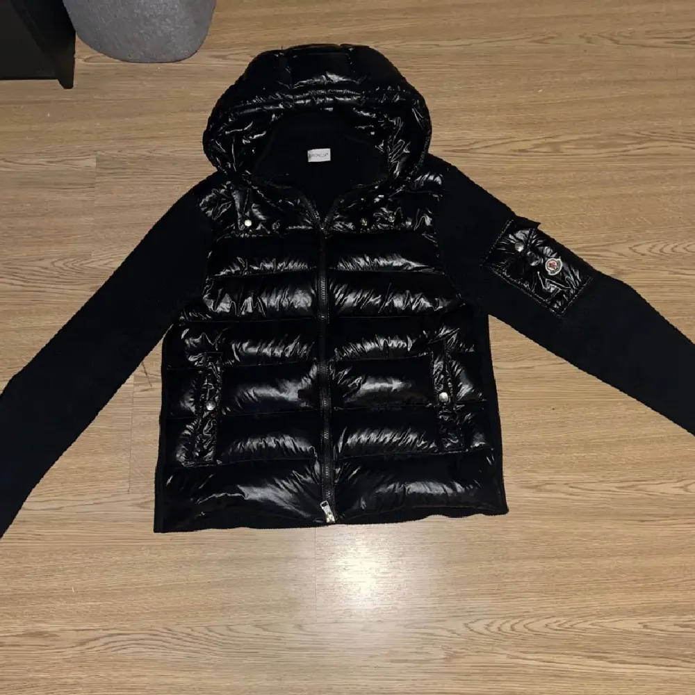 Säljer en snygg svart pufferjacka från Moncler. Jackan har en glansig finish med quiltade paneler och en dragkedja framtill. Den har en huva och långa ärmar, perfekt för kyliga dagar. Insidan är mjuk och bekväm, vilket gör den idealisk för både höst . Jackor.