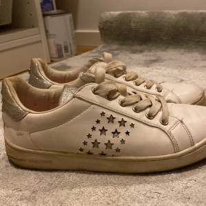 Säljer ett par vita sneakers med coola stjärndetaljer på sidan. Skorna har en glittrig silverdetalj vid hälen och är i välanvänt skick. Perfekta för en avslappnad stil med en touch av glitter!