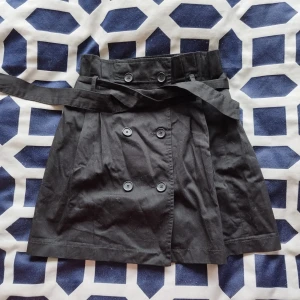 Svart kjol med knappar och bälte - Säljer en snygg svart kjol med knappar framtill och ett matchande bälte. Kjolen har en stilren design med dubbelknäppning och är perfekt för både vardag och fest. Materialet känns slitstarkt och bekvämt.