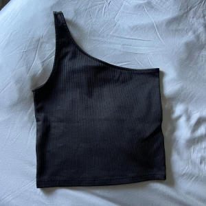 Svart ribbat one shoulder linne - Ribbar one shoulder linne, för litet för mig men i nyskick! Uppvikt på andra bilden då jag hade en högre kjol, går ner förbi naveln i vanliga fall!