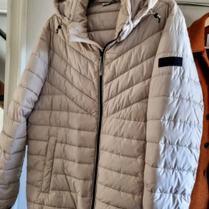 Beige pufferjacka från Tom Tailor - Säljer en snygg och bekväm beige pufferjacka från Tom Tailor. Jackan har en dragkedja framtill och en praktisk huva. Perfekt för kyliga dagar och håller dig varm och stilren. Passar bra till både vardag och utflykter!