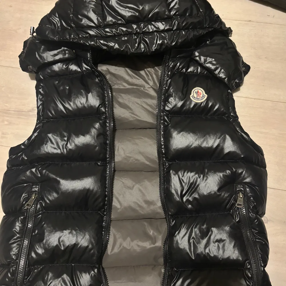 Säljer en snygg och glansig svart dunväst från Moncler. Västen har en quiltad design med dragkedja framtill och två praktiska fickor med dragkedjor. Perfekt för kyliga dagar när du vill hålla stilen. Moncler-loggan är broderad på bröstet. Passar både till vardags och lite finare tillfällen.. Jackor.