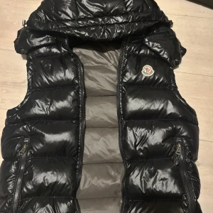 Svart dunväst från Moncler - Säljer en snygg och glansig svart dunväst från Moncler. Västen har en quiltad design med dragkedja framtill och två praktiska fickor med dragkedjor. Perfekt för kyliga dagar när du vill hålla stilen. Moncler-loggan är broderad på bröstet. Passar både till vardags och lite finare tillfällen.