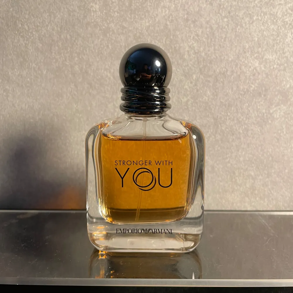 Säljer en flaska Stronger With You Eau de Toilette från Emporio Armani. Den har en elegant och stilren design med en rundad svart kork och en klar glasflaska som visar den gyllene doften inuti. Doften har en varm och söt karaktär med inslag av vanilj, balsamico och nötiga toner. Perfekt för den som vill ha en aromatisk och pudrig doft med en hint av örtighet.. Parfym.