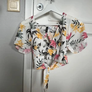 Blommig topp  - Blommig topp med urringning och öppen rygg. Armarna är lösa och öppna. Knyts baktill vilket ger en korsad rygg med knytning. Magkort och urringad. Bekväm och fin.