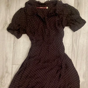Mörkbrun prickig klänning Indiska - Säljer en superfin mörkbrun klänning med små vita prickar från Indiska. Den har korta ärmar och en söt volangdetalj längst ner. Perfekt för både vardag och fest!