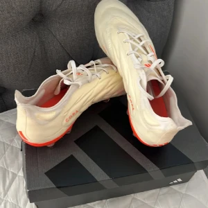 Elite copas addidas - Säljer ett par snygga fotbollsskor från Adidas i vitt med coola orange detaljer. Skorna har snörning och är i nyskick, perfekta för fotbollsmatcher. De har en modern design med de klassiska tre ränderna på sidan. Sulan är utrustad med dobbar för optimalt grepp på planen.