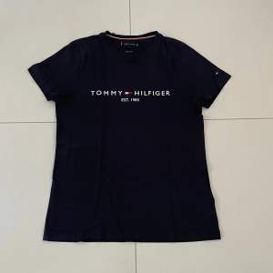 Säljer en stilren mörkblå t-shirt från Tommy Hilfiger med deras ikoniska logga och 'EST. 1985' tryckt på framsidan. Perfekt för en casual look eller att matcha med jeans. T-shirten har korta ärmar och är i bra skick.