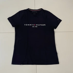 Svart t-shirt från Tommy Hilfiger - Säljer en stilren mörkblå t-shirt från Tommy Hilfiger med deras ikoniska logga och 'EST. 1985' tryckt på framsidan. Perfekt för en casual look eller att matcha med jeans. T-shirten har korta ärmar och är i bra skick.