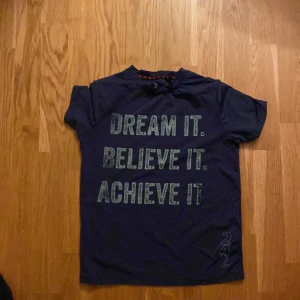 Mörkblå t-shirt med motiverande text - Säljer en mörkblå t-shirt med korta ärmar och motiverande texten 'Dream it. Believe it. Achieve it.' i ljusare blått. Perfekt för vardagsbruk eller träning. T-shirten är i bra skick och passar till många olika outfits.