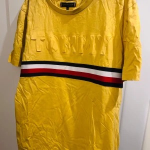 Gul t-shirt från Tommy Hilfiger - Säljer en snygg gul t-shirt från Tommy Hilfiger i ekologisk bomull. Den har ett coolt tryck med 'TOMMY' i relief och en randig detalj i rött, vitt och blått över bröstet. Har kortärmade design. Har en liten brun fläck som ev går att tvätta bort.