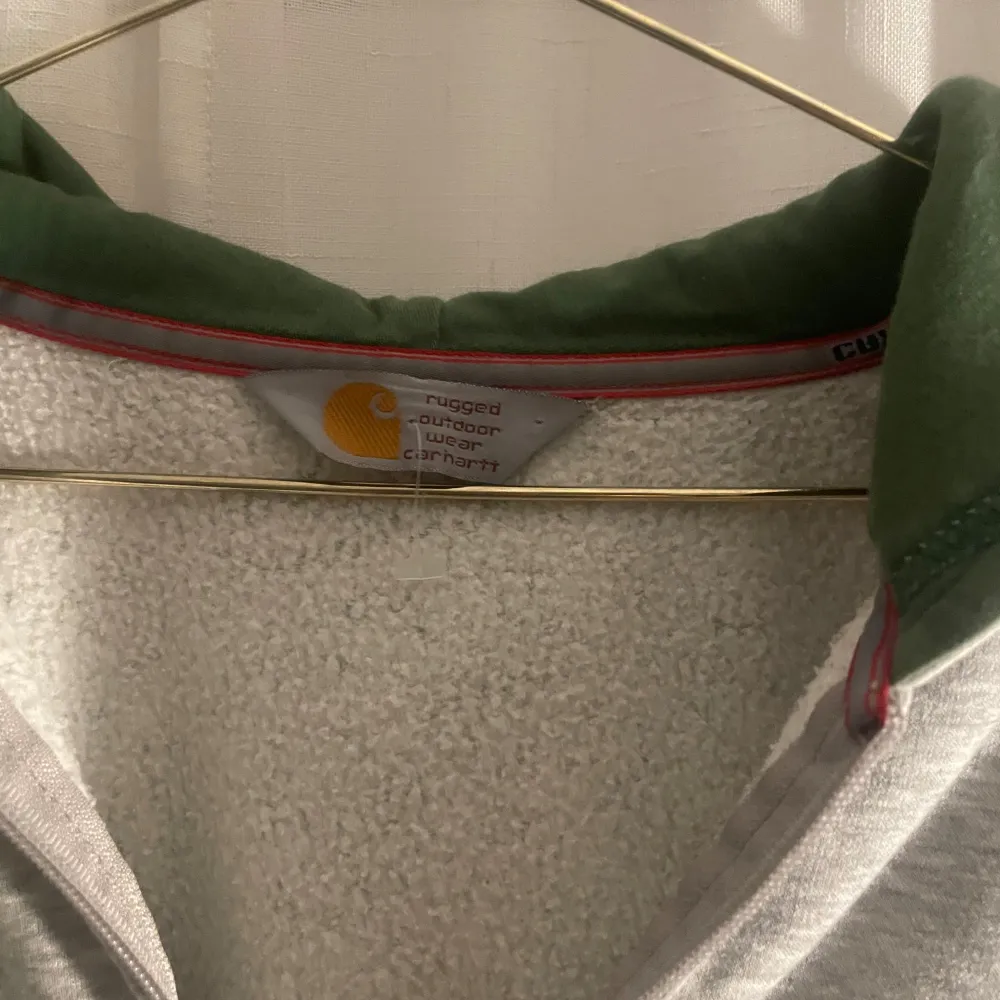 Säljer en grå hoodie från Carhartt med dragkedja och en mysig grön insida i luvan. Den har en klassisk passform med ribbade muddar och fickor framtill. Superbekväm och stilren!. Neuletakit & Villapaidat.