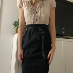 Beige och svart klänning från H&M - Säljer en elegant knälång klänning från H&M i storlek M, passar även small (som jag har). Överdelen är beige/rosa med fina volangdetaljer och knappar framtill, medan kjolen är svart och knyts i midjan för en smickrande passform 
