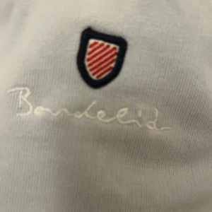 Vit tröja från Bardeid - Säljer en stilren vit tröja från Bardeid. Den har en enkel design med ett litet broderat emblem på bröstet. Tröjan är långärmad och perfekt för en avslappnad look. Den är i bra skick med en liten fläck på framsidan. Perfekt för både vardag och chillhäng!