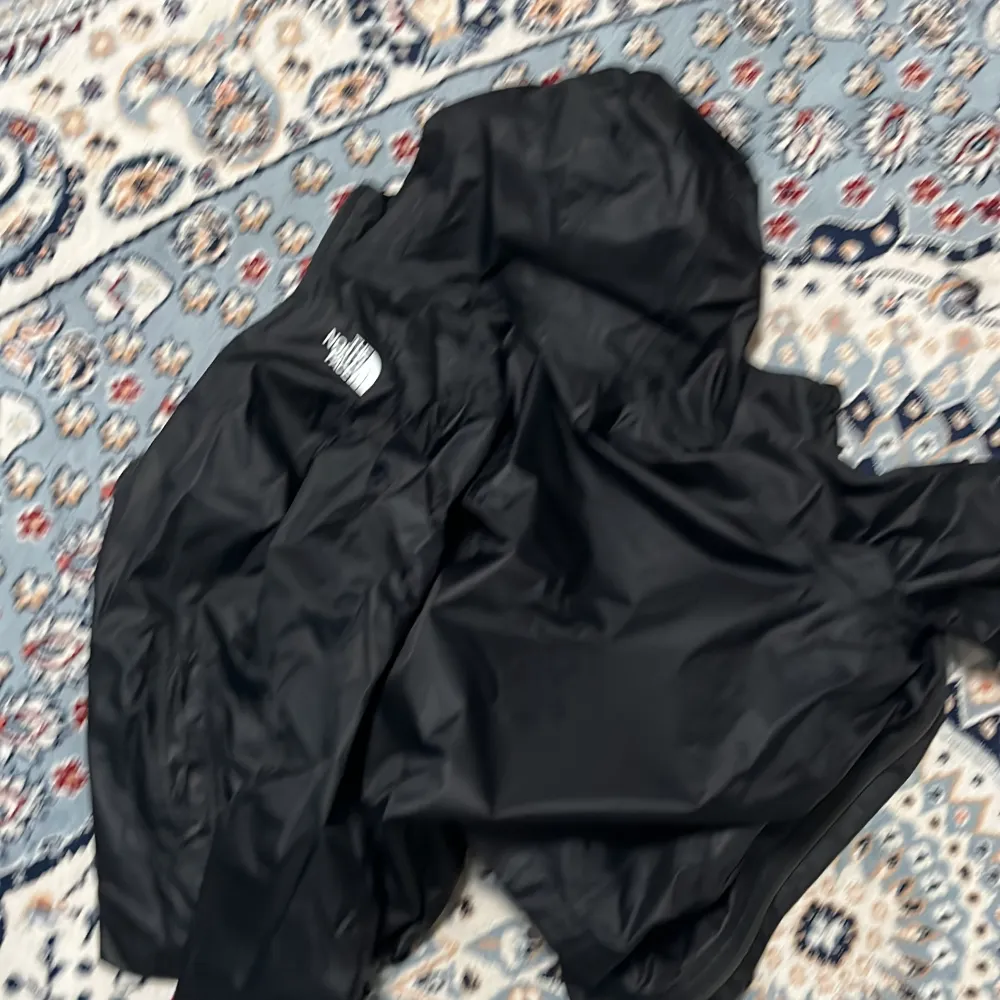 Säljer en svart vindjacka från The North Face. Jackan är perfekt för blåsiga dagar och har en stilren design med The North Face-loggan på bröstet. Den är lätt och smidig att bära med sig. Passar bra för både vardag och utomhusaktiviteter.. Jackor.