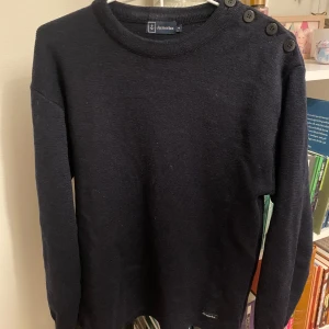 Svart stickad tröja från Armor Lux - Säljer en stilren svart stickad tröja från Armor Lux. Den har långa ärmar och snygga knappar på axeln som ger en marin känsla. Perfekt för både höst och vinter, och den är supermjuk och bekväm. Passar till alla tillfällen!