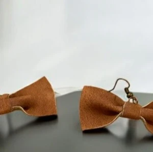 Bruna rosettörhängen - Charmiga örhängen i form av bruna rosetter i konstläder. Perfekta för att ge en lekfull touch till din outfit. De har en enkel krok för enkel användning.