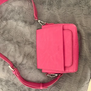 Rosa axelväska i syntet - Snygg rosa axelväska i syntetmaterial med krokodilmönster. Väskan har en justerbar axelrem och en stilren design med ett lock framtill. Perfekt för att ge en färgklick till din outfit.