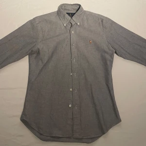 Grå skjorta från Ralph Lauren - Snygg grå skjorta från Ralph Lauren i slim fit. Skjortan har långa ärmar och knappar framtill. Perfekt för en stilren look med broderad logga på bröstet. Skjortan är varsamt använd och har inga defekter. 