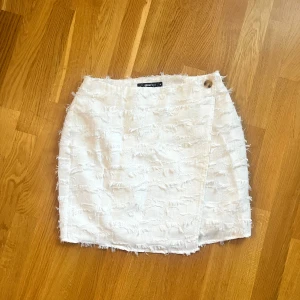 Vit omlottkjol med fransar - Snygg vit omlottkjol från Gina med fransiga detaljer och en knapp i midjan. Perfekt för en stilren look.