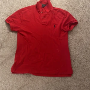 Röd kortärmad pikétröja från Ralph Lauren - Snygg röd pikétröja från Ralph Lauren i slim fit. Tröjan har en klassisk krage med knappar och det ikoniska broderade logotypen på bröstet. Perfekt för en stilren look.