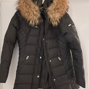 Svart dunjacka från Rockandblue - Snygg svart dunjacka från Rockandblue med quiltad design och pälskantad huva. Jackan har dragkedjor på fickorna och en cool patch på ärmen. Perfekt för kalla dagar när du vill hålla stilen. 🖤