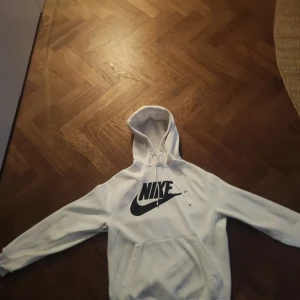 Nike hoodie - Säljer en stilren vit hoodie från Nike med det klassiska logotyptrycket i svart på framsidan. Hoodien har en känguruficka och justerbar huva med snören. Perfekt för en avslappnad stil.