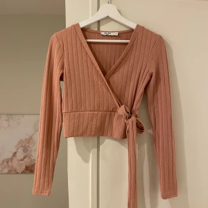 Rosa ribbad omlottopp från NA-KD - Säljer en snygg rosa ribbad omlottopp från NA-KD. Toppen har långa ärmar och en knytning i midjan som ger en fin passform. Perfekt för en stilren look.