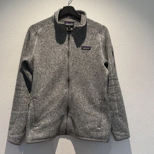Grå fleecejacka från Patagonia - Säljer en stilren grå fleecejacka från Patagonia med dragkedja framtill. Jackan har en normal passform och är perfekt för kyligare dagar. Den har en broderad logotyp på bröstet och praktiska fickor med dragkedja.