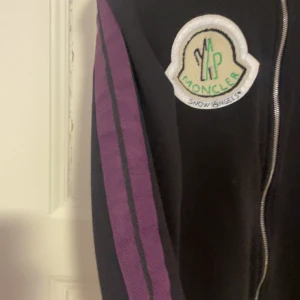 Svart jacka från Moncler x Palm Angels - Snygg svart jacka från Moncler x Palm Angels med dragkedja och coola detaljer. Har ett broderat märke på bröstet och texten 'Mind Control' på ryggen. Lila ränder längs ärmarna ger en extra touch.