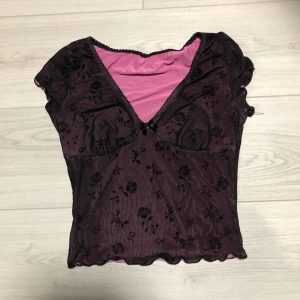 Svart blommig topp med rosa foder - Snygg svart topp med blommigt mönster och rosa foder. Toppen har en v-ringad design med en liten rosett framtill. Den är kortärmad och har en fin volangdetalj vid ärmslut och nederkanten 💕
