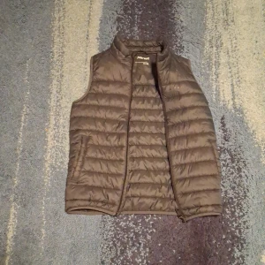 Olive dunväst från Everest - Säljer en brun dunväst från Everest. Västen har en dragkedja framtill och är quiltad för extra värme. Perfekt för kyliga dagar när du vill hålla dig varm utan att bära en jacka. Passar bra för både vardag och utomhusaktiviteter.