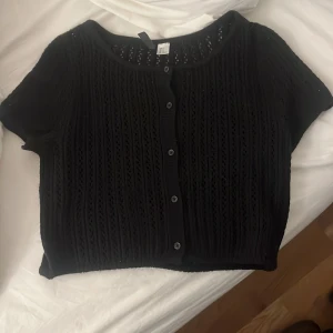 Svart kortärmad kofta från H&M - Säljer en svart kortärmad kofta från H&M:s Divided-kollektion. Koftan har ett fint stickat mönster och knappar framtill. Perfekt för lager-på-lager-stil eller som ett lättare plagg under svalare dagar.