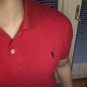 Röd pikétröja från Ralph Lauren - Säljer en klassisk röd pikétröja från Ralph Lauren med den ikoniska loggan broderad på bröstet. Tröjan har en krage och knappar framtill, perfekt för en stilren look. Säljer pga att jag inte använder 