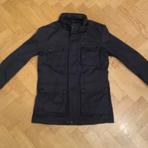 Fieldjacket - Snygg blå fieldjacket med flera fickor framtill. Perfekt för höst och vår med sin stilrena design och praktiska detaljer. Jackan har långa ärmar och en hög krage för extra skydd mot väder och vind. skick 9/10 knappt använd 